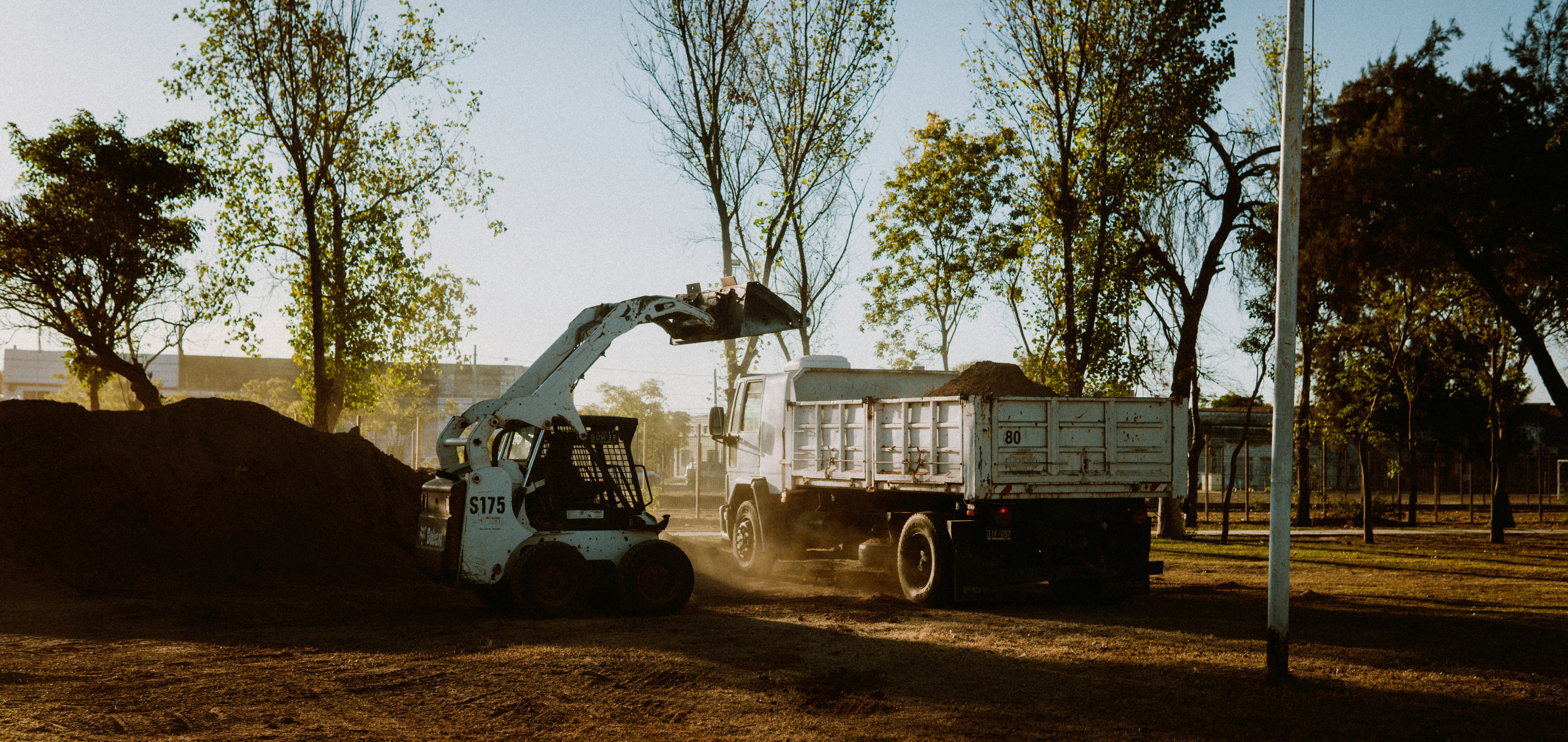 Excavator loading soil onto a dump truck at a construction site with trees in the background.